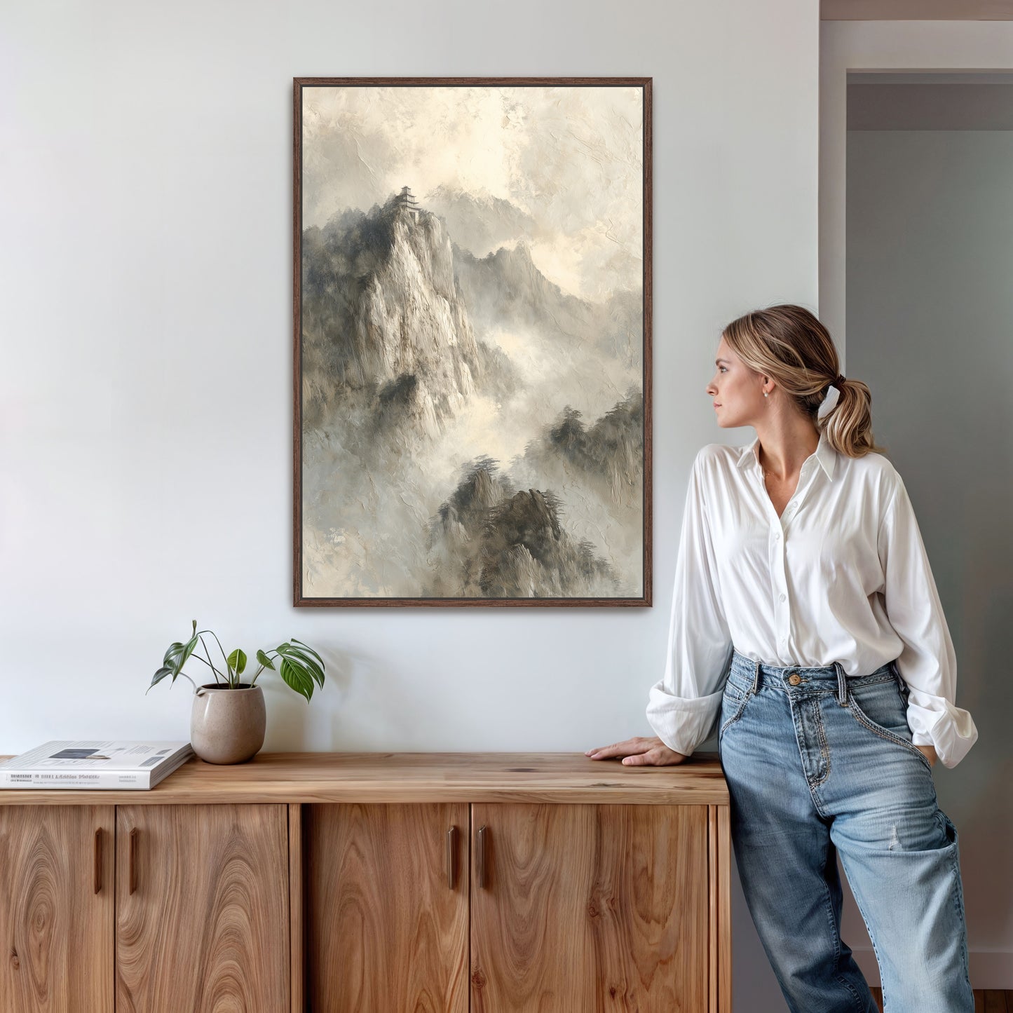 A woman in a white blouse and blue jeans stands in a room, leaning against a wooden cabinet, with a framed landscape painting hanging on the wall above her.