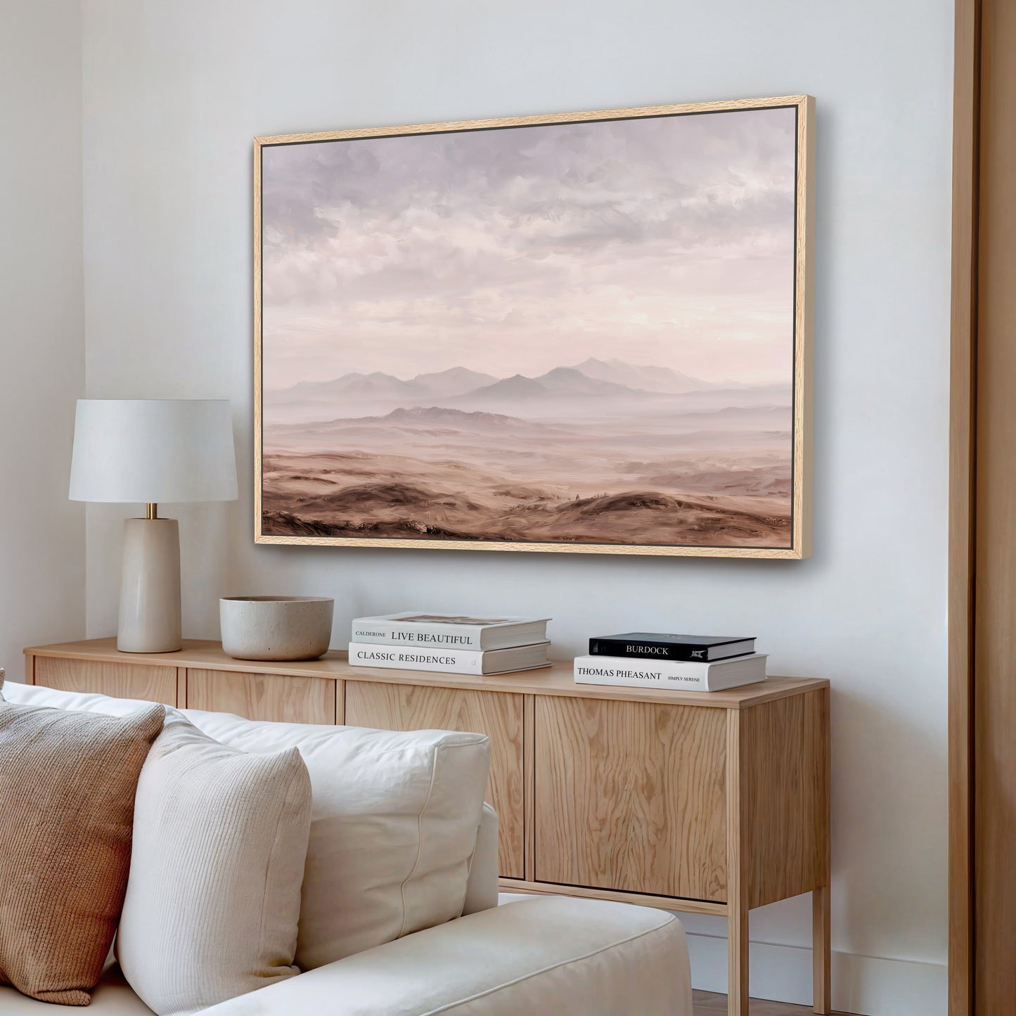 a living room with a large framed landscape painting hanging on the wall above a wooden cabinet. The painting depicts a desert scene with mountains in the background. The cabinet has a lamp and some books on it, creating a cozy and inviting atmosphere.