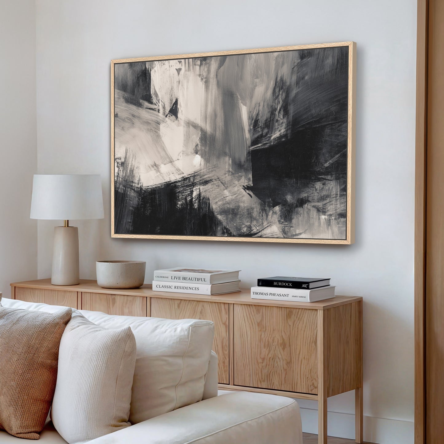 a living room with a large abstract painting hanging on the wall above a wooden cabinet. The cabinet has a white lamp on top and a stack of books. The room has a white sofa and a wooden coffee table.