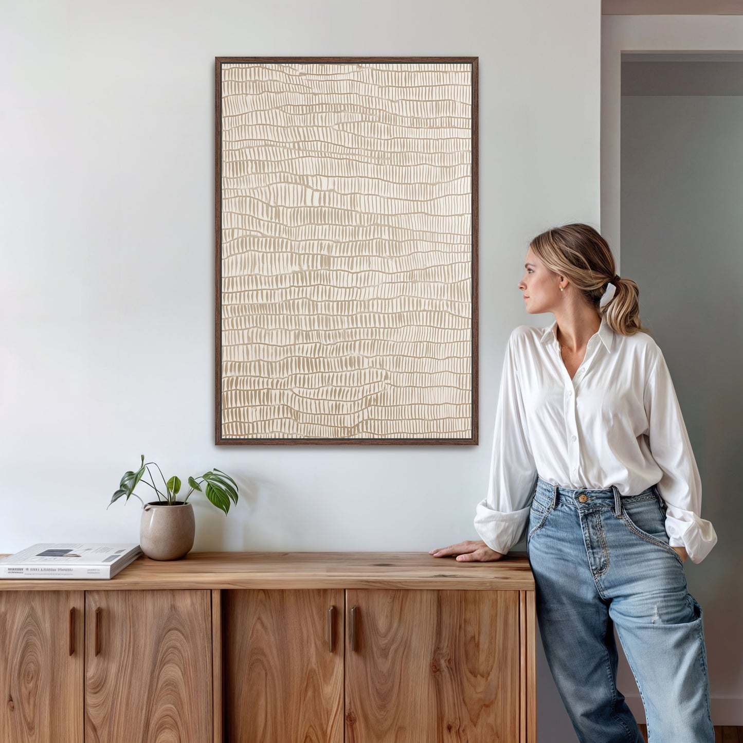 A woman in a white blouse and blue jeans stands next to a wooden cabinet, leaning against it with her hand on her hip. The cabinet has a plant on top of it, and there is a framed picture hanging on the wall above it.