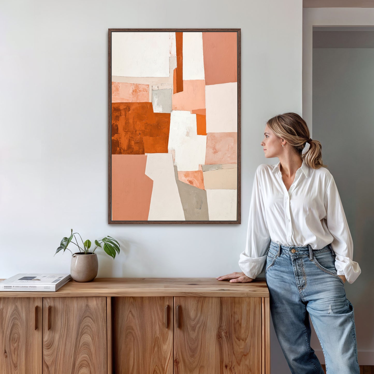 A woman in a white shirt and blue jeans stands in a room, leaning against a wooden cabinet, with a framed abstract painting hanging on the wall above her.