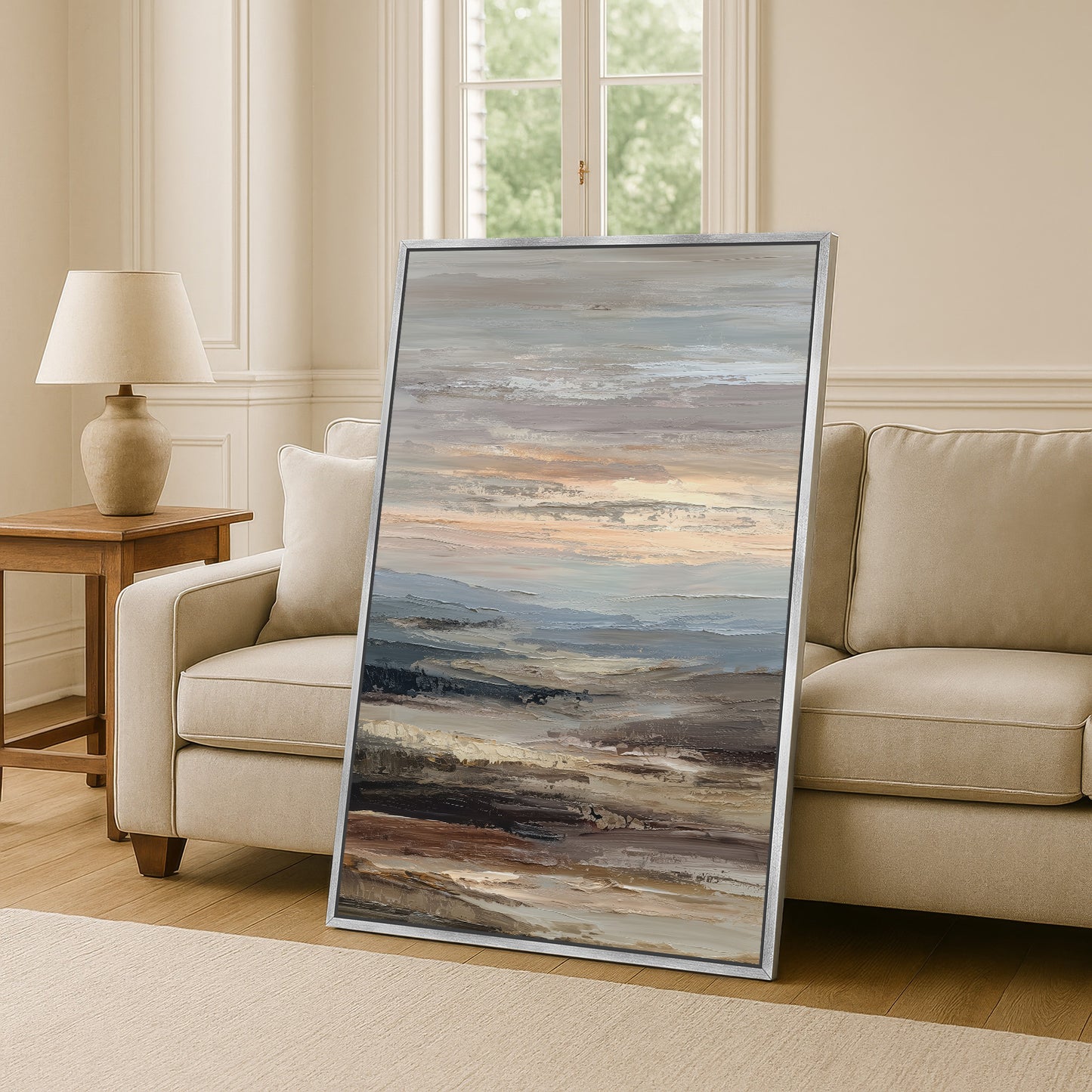 a large, framed painting of a landscape depicting a serene, cloudy sky with rolling hills and mountains in the background. The painting is placed on a wooden floor in a living room setting, with a beige sofa and a wooden side table nearby.