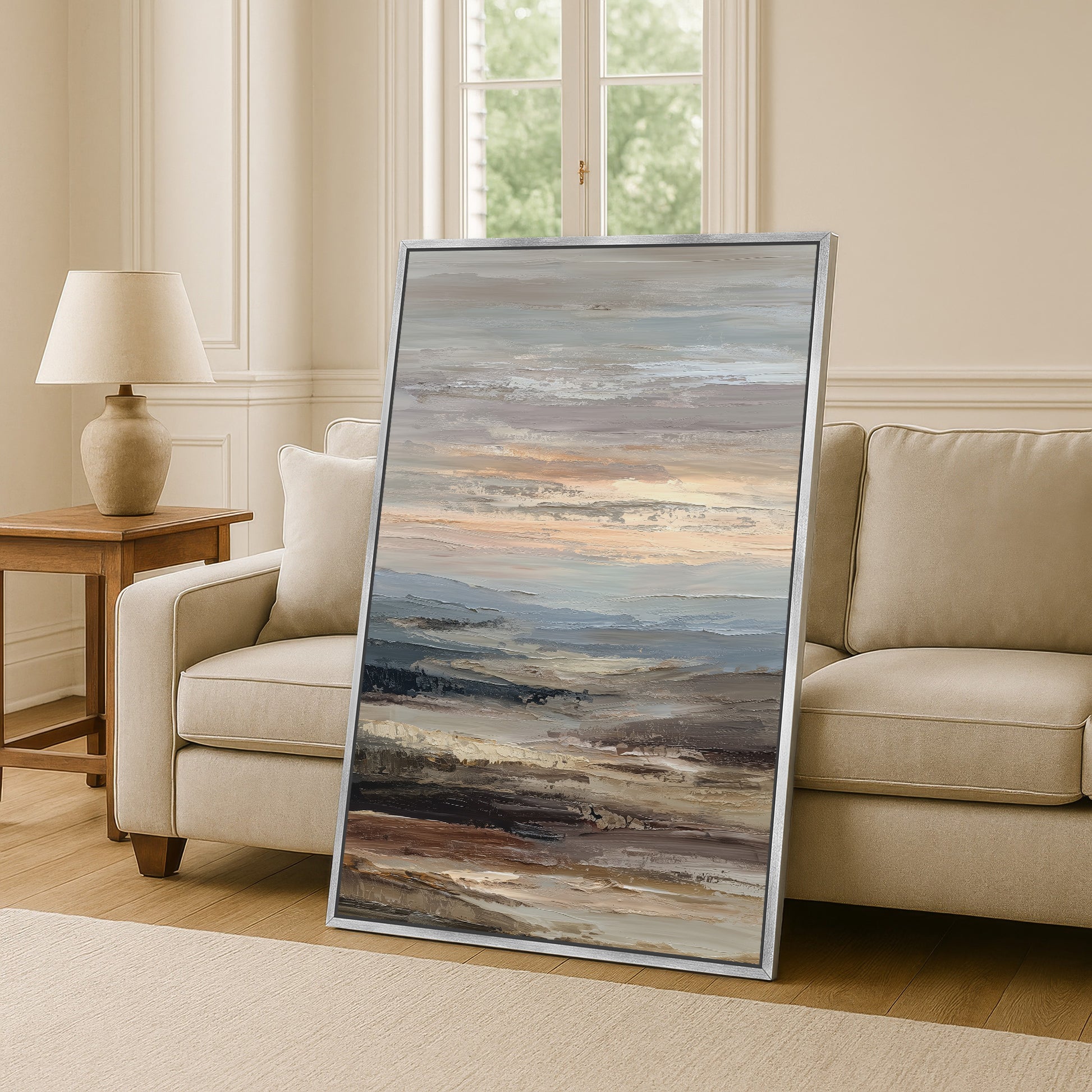 a large, framed painting of a landscape depicting a serene, cloudy sky with rolling hills and mountains in the background. The painting is placed on a wooden floor in a living room setting, with a beige sofa and a wooden side table nearby.