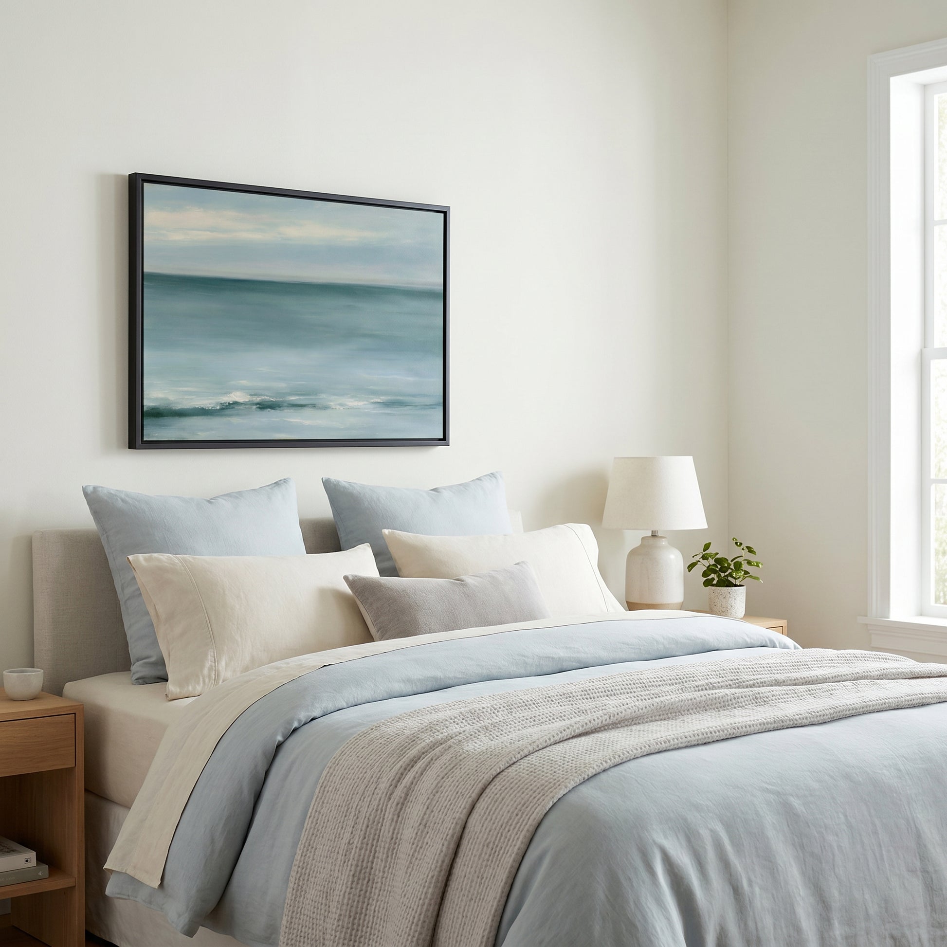 a serene bedroom scene with a large bed adorned with a blue and gray comforter, a white pillow, and a gray throw blanket. Above the bed, a framed painting of a tranquil ocean landscape is displayed on the wall. The room is well-lit, with a window on the right side allowing natural light to enter the space.