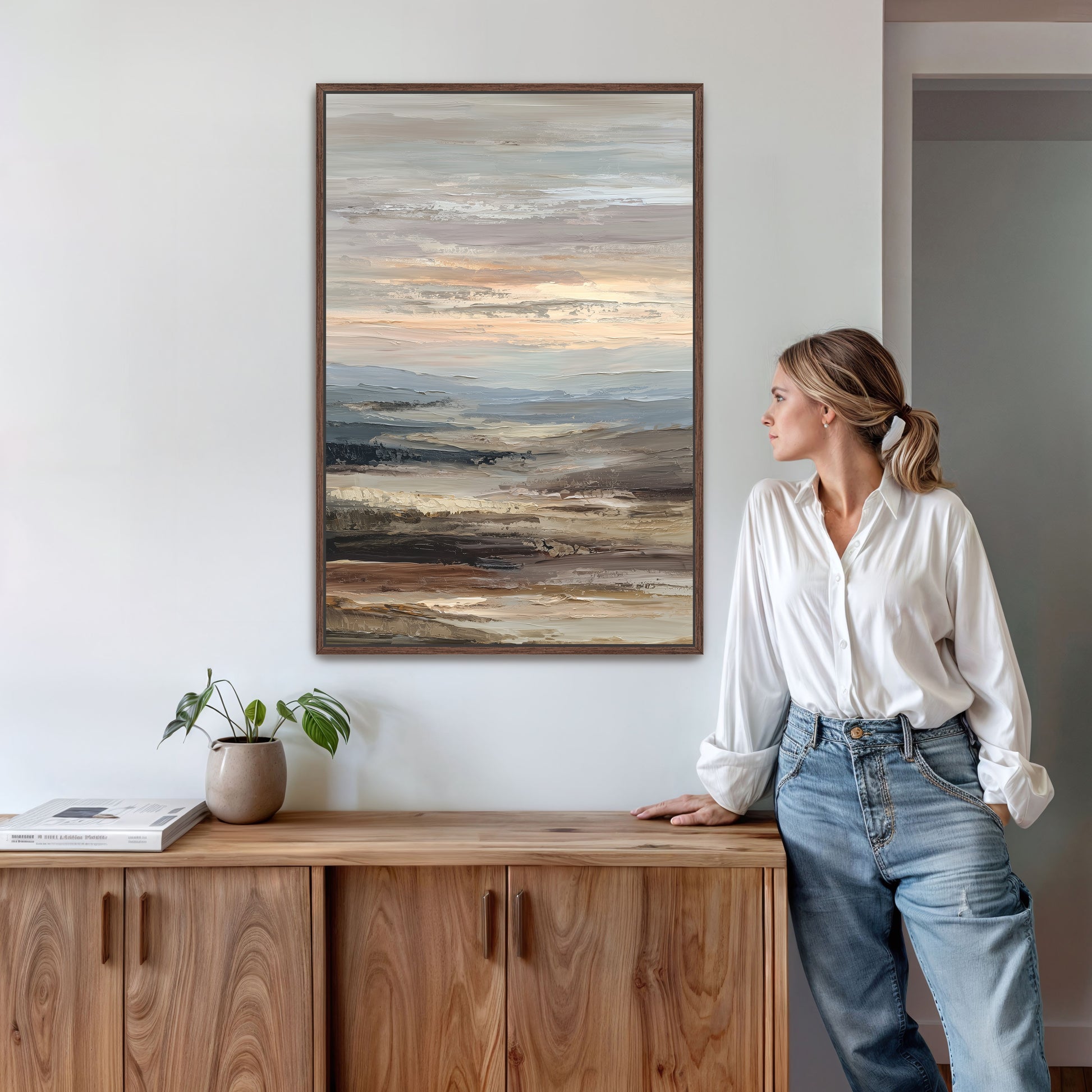 A woman in a white blouse and blue jeans stands next to a wooden cabinet, leaning against it and looking to the side. Above her, a large painting of a landscape is hanging on the wall.