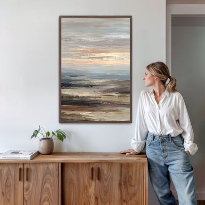 A woman in a white blouse and blue jeans stands next to a wooden cabinet, leaning against it and looking to the side. Above her, a large painting of a landscape is hanging on the wall.
