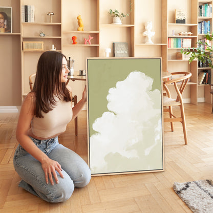 Woman kneeling beside framed Rise Canvas Wall Art featuring a cloud against muted sage background in a modern living room