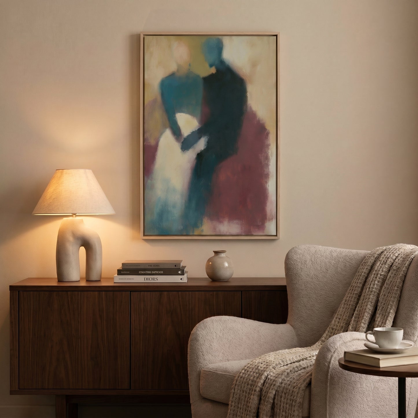 a cozy living room with a painting of two people on the wall, a wooden sideboard with a lamp and books, a beige armchair, and a small table with a coffee cup and saucer.