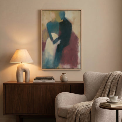 a cozy living room with a painting of two people on the wall, a wooden sideboard with a lamp and books, a beige armchair, and a small table with a coffee cup and saucer.