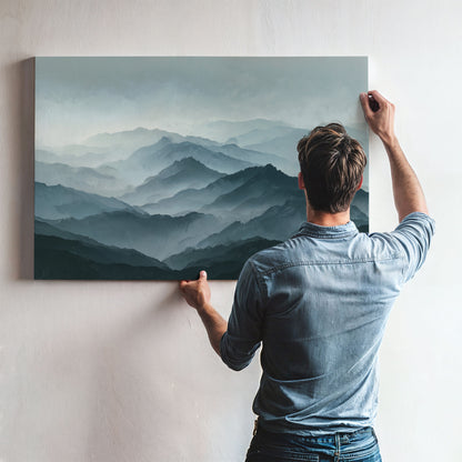 A man is hanging a large landscape painting on a wall, which depicts a mountainous scene with a hazy, misty atmosphere.