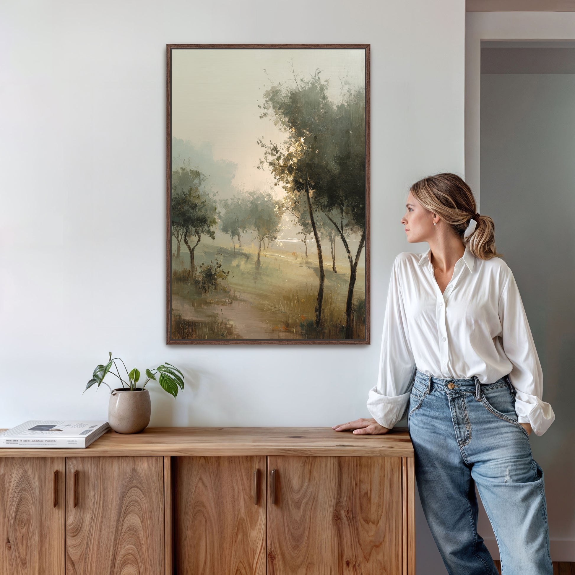 A woman in a white blouse and blue jeans stands in a room, leaning against a wooden cabinet, with a framed landscape painting hanging on the wall above her.