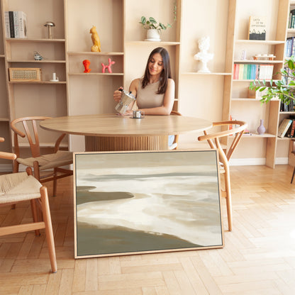 Emie Canvas Print framed coastal landscape with soft beige, ivory, and olive tones displayed on the floor beside a dining table in a modern room
