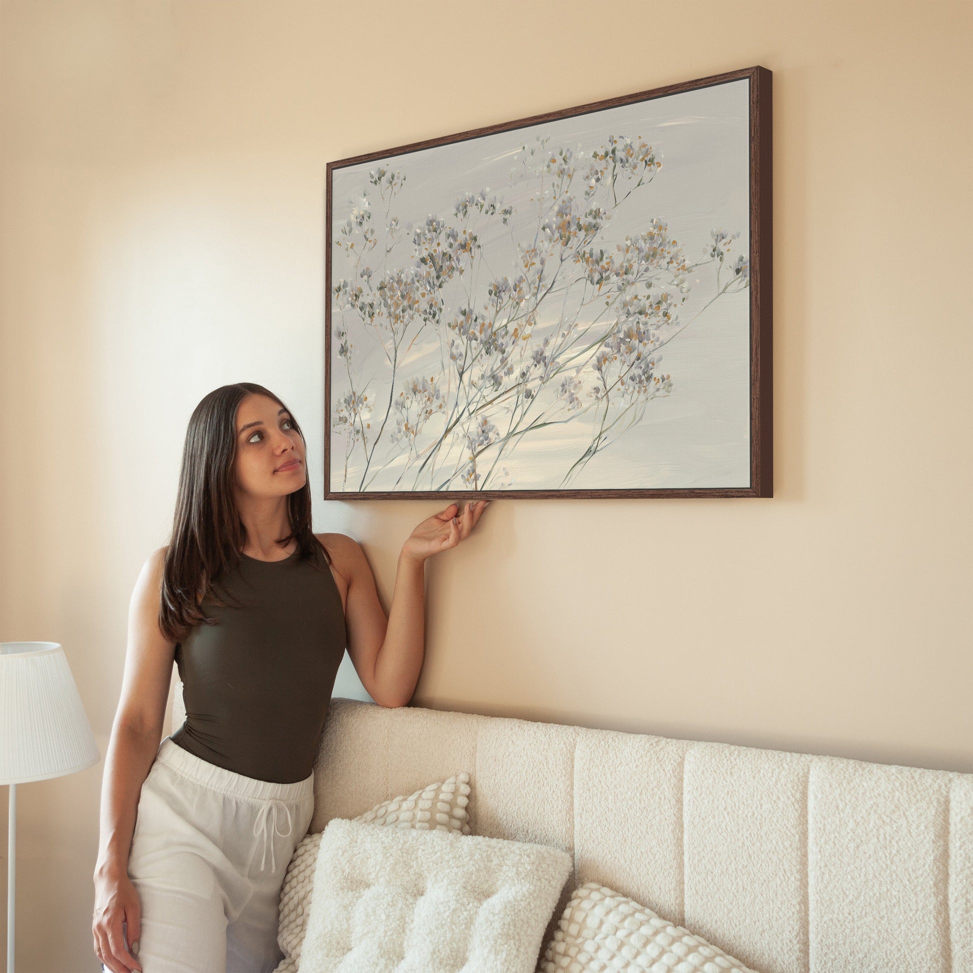 Woman admiring framed Breeze Wall Art featuring delicate wildflowers on a neutral wall above a beige couch.