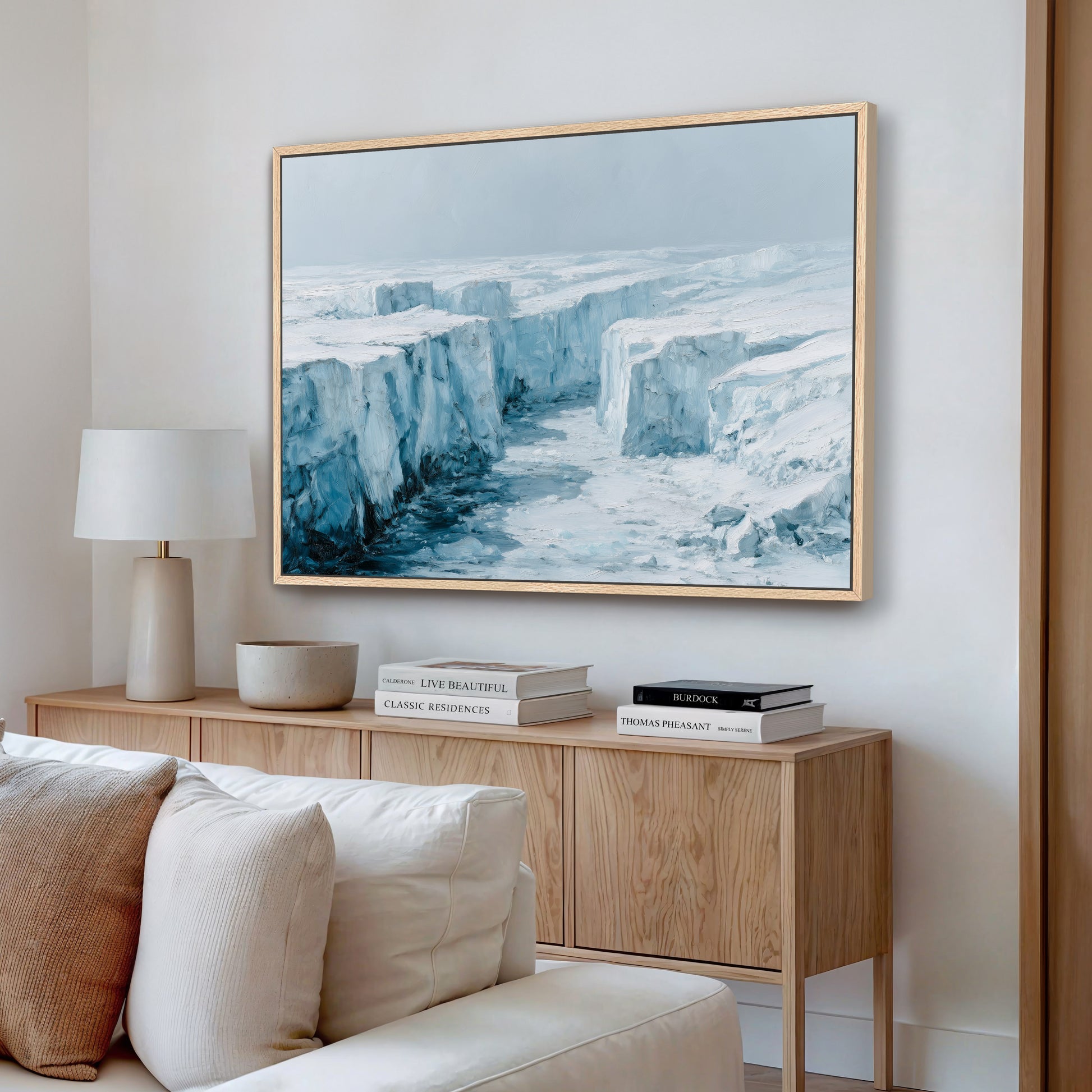 a large framed painting of a snowy, icy landscape hanging on a wall above a wooden cabinet with books and a lamp.