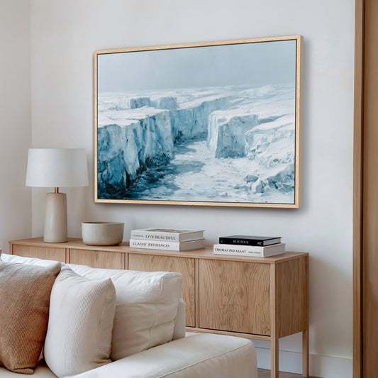 a large framed painting of a snowy, icy landscape hanging on a wall above a wooden cabinet with books and a lamp.