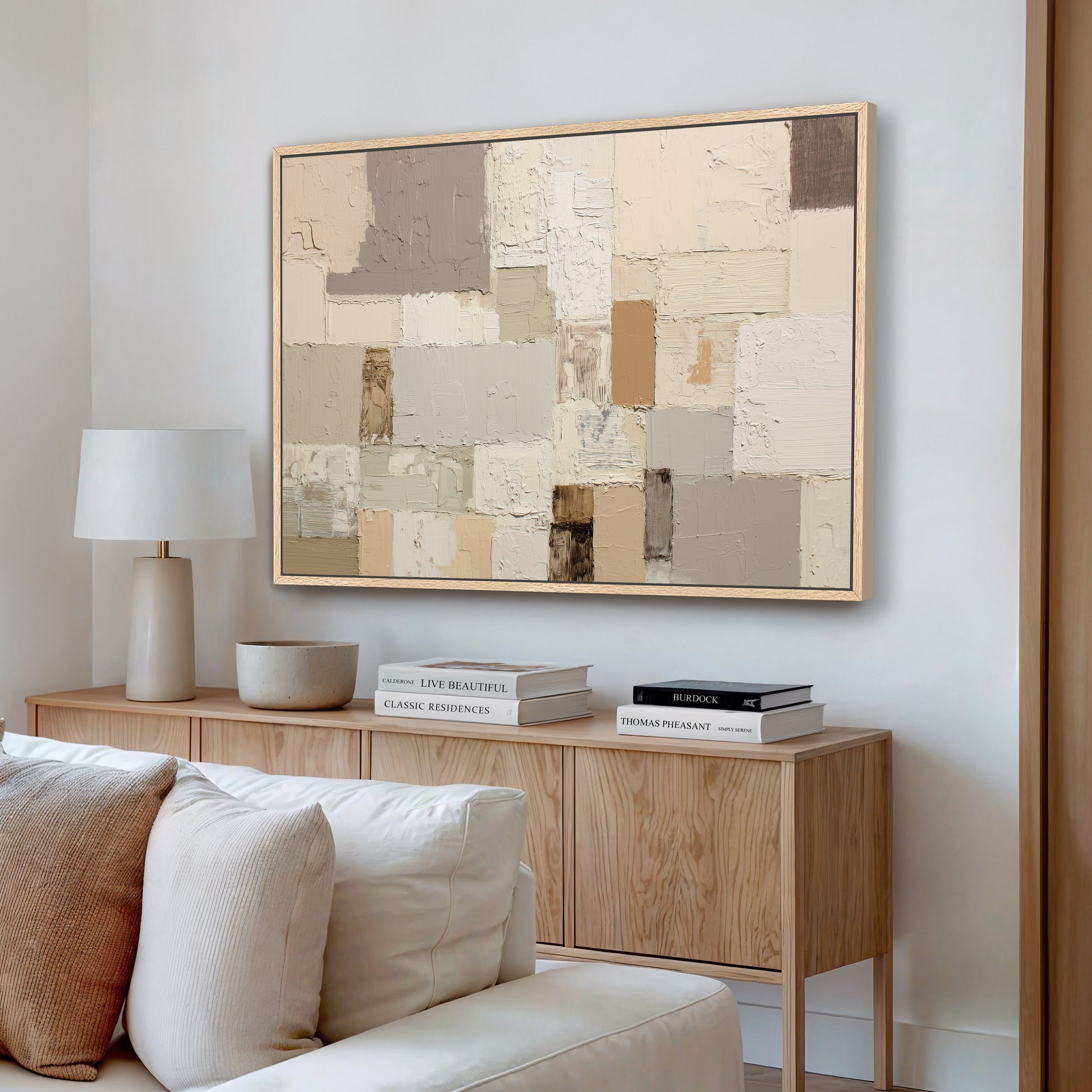 a living room with a large abstract painting hanging on the wall above a wooden cabinet. The cabinet has a bowl and two books on top of it. The room also includes a white couch and a lamp.