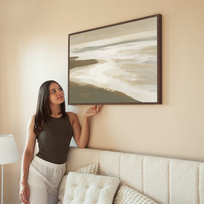 Woman hanging Emie canvas print of tranquil coastal landscape in neutral tones on beige wall above couch