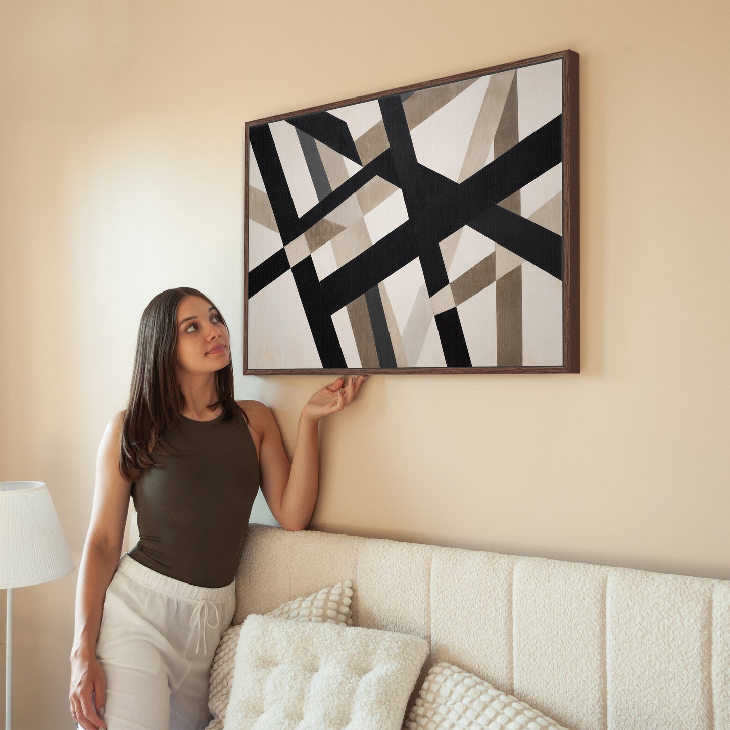 Axis canvas print with bold geometric black and neutral intersecting lines in a brown wooden frame hanging on a beige wall above a sofa with a woman admiring it