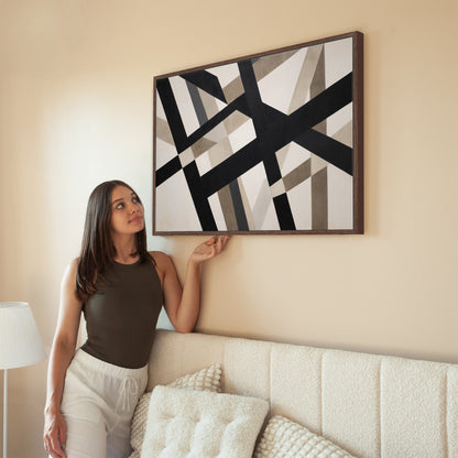 Axis canvas print with bold geometric black and neutral intersecting lines in a brown wooden frame hanging on a beige wall above a sofa with a woman admiring it