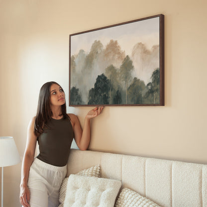 Woman admiring Fable Canvas Print of misty treetops in muted olive and ivory tones hung above beige sofa