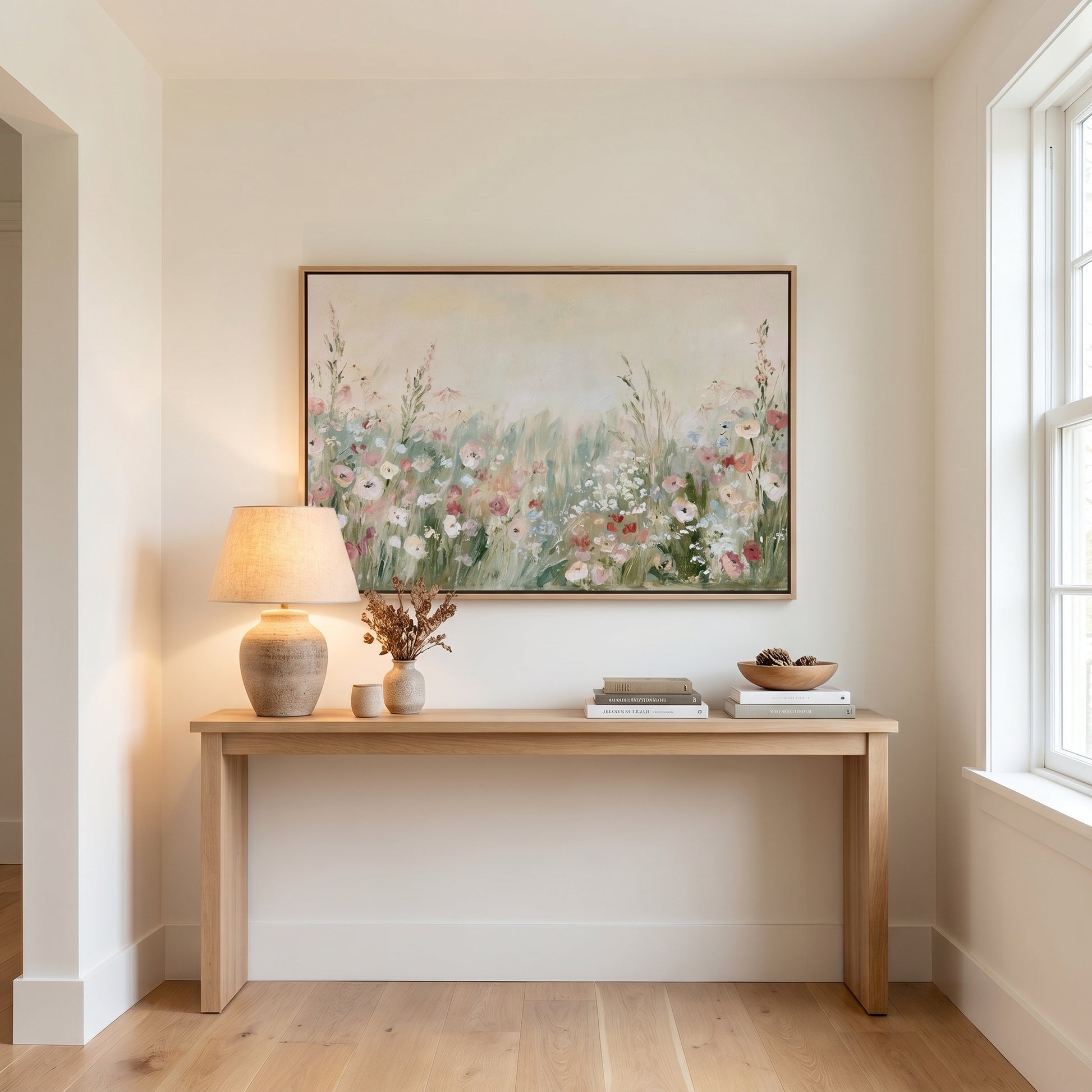 a wooden console table with a lamp, a vase of flowers, and a stack of books on it, set against a white wall with a large painting hanging above it.