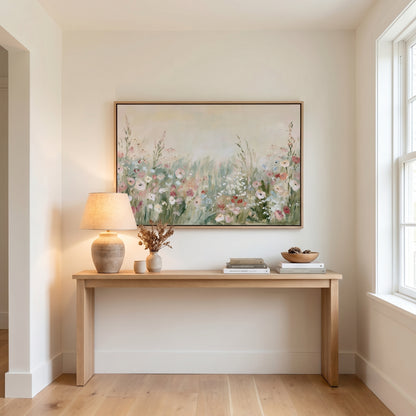 a wooden console table with a lamp, a vase of flowers, and a stack of books on it, set against a white wall with a large painting hanging above it.