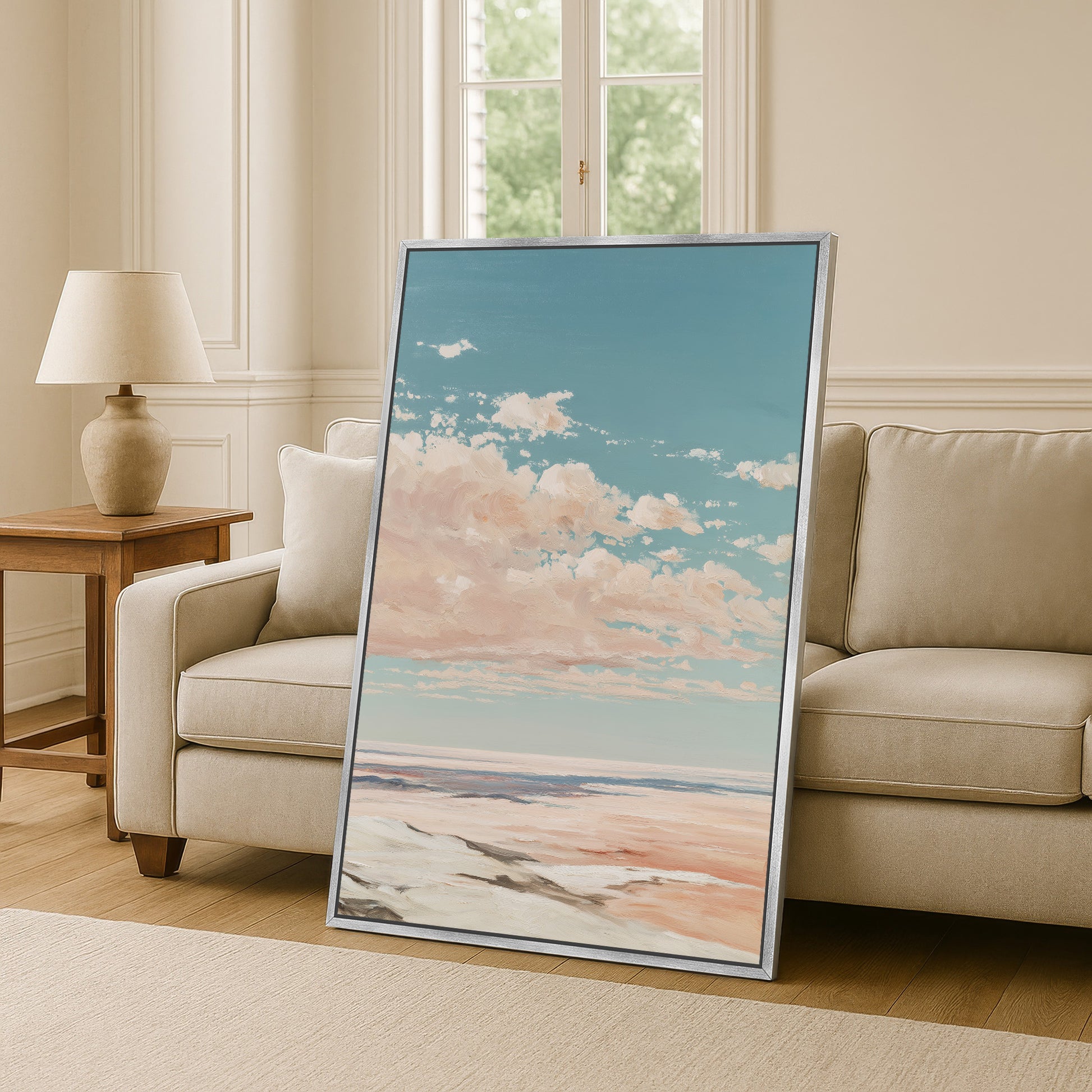 a large framed painting of a serene landscape with a blue sky, fluffy clouds, and a sandy beach in the background, placed on a wooden floor in a living room setting.