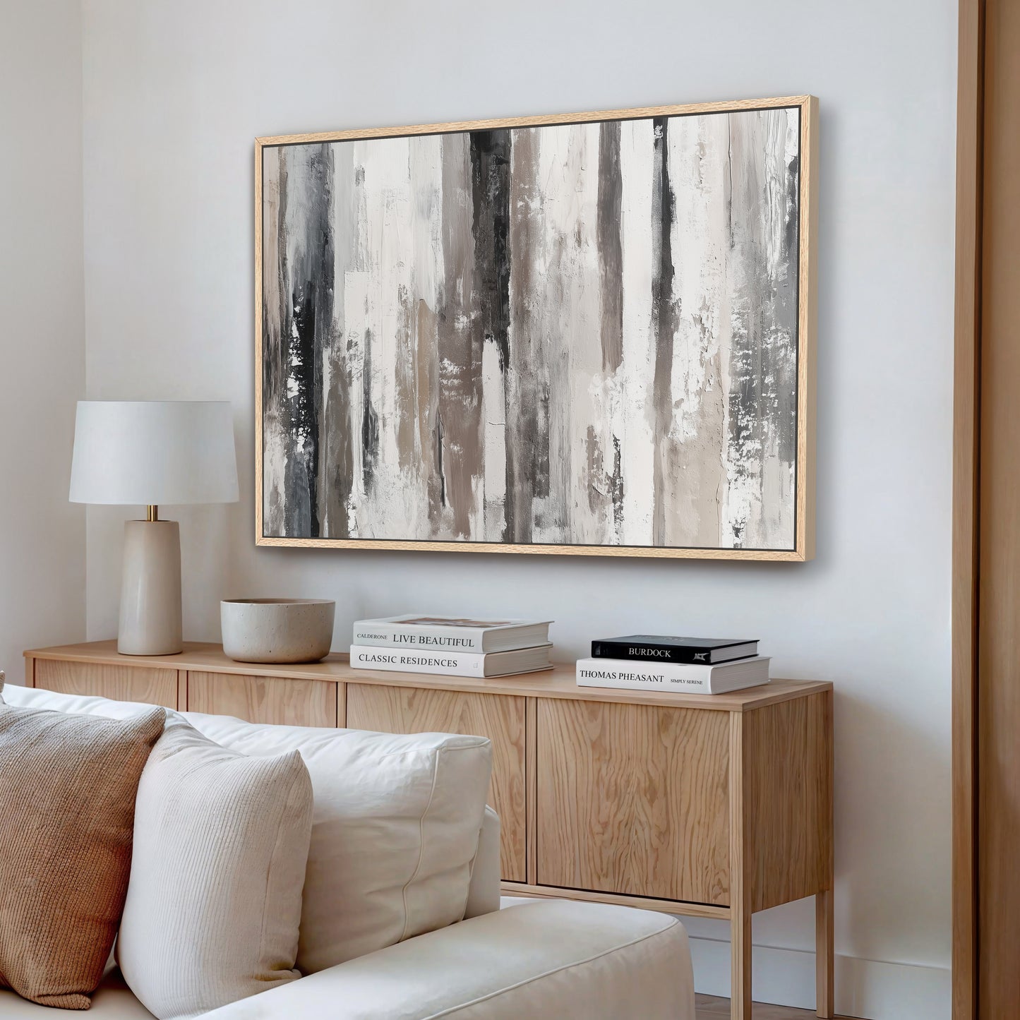 a modern living room with a large abstract painting hanging on the wall above a wooden console table. The console table has a lamp and a stack of books on it. The room also includes a white sofa and a wooden bookshelf.
