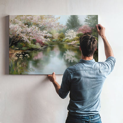 A man is hanging a large painting of a serene landscape on a wall, which features a tranquil pond surrounded by blooming trees.