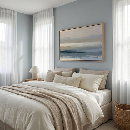 a serene and cozy bedroom with a large bed adorned with white linens and beige throw pillows. Above the bed, a large painting of a landscape with a blue sky and water adds a touch of nature to the room. The room is illuminated by natural light streaming in through the windows, creating a warm and inviting atmosphere.