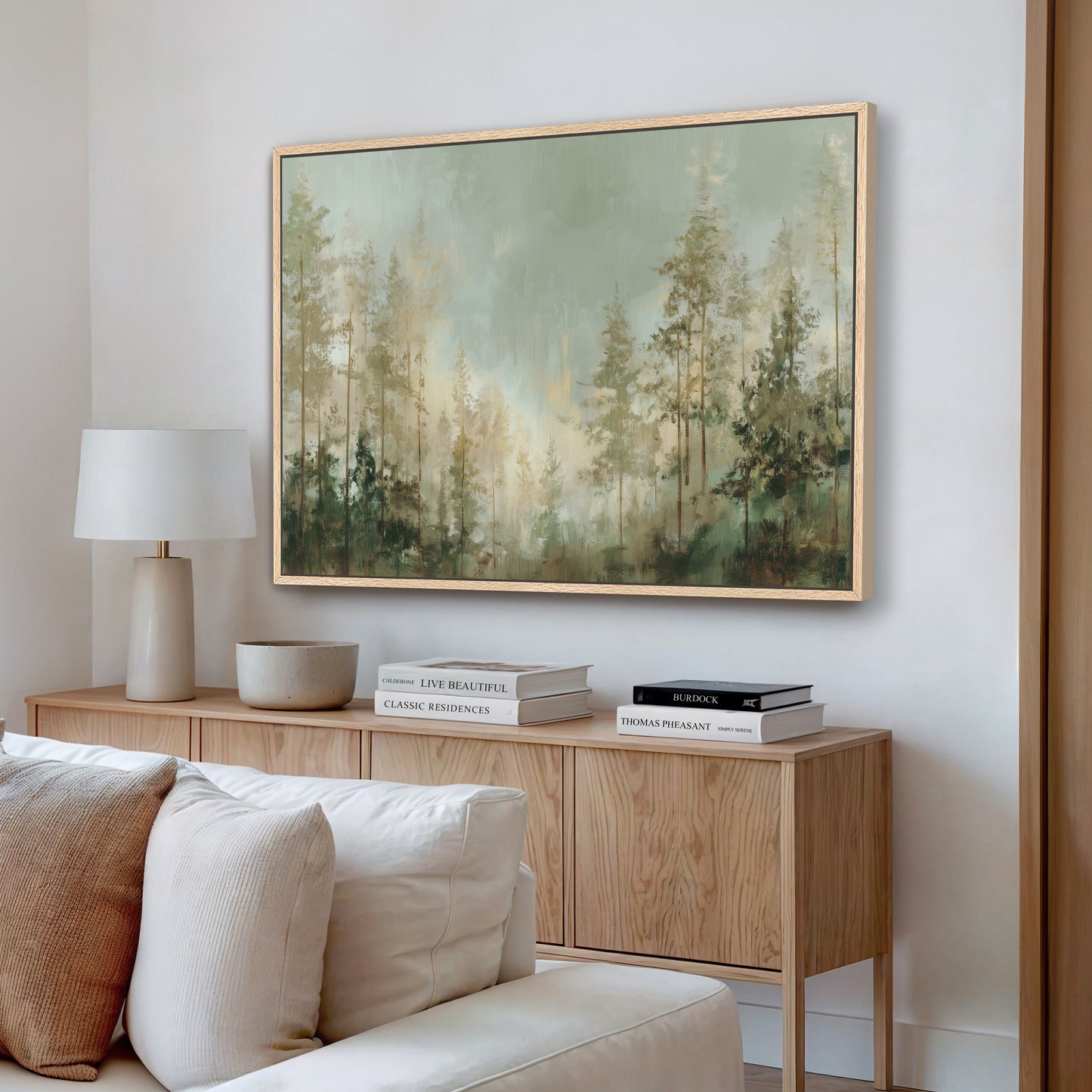a living room with a large painting of a forest scene hanging on the wall above a wooden cabinet. The cabinet has a bowl and a stack of books on top. There is a white couch in the room, and the overall color scheme is neutral, with the painting and the wooden cabinet providing a warm contrast.