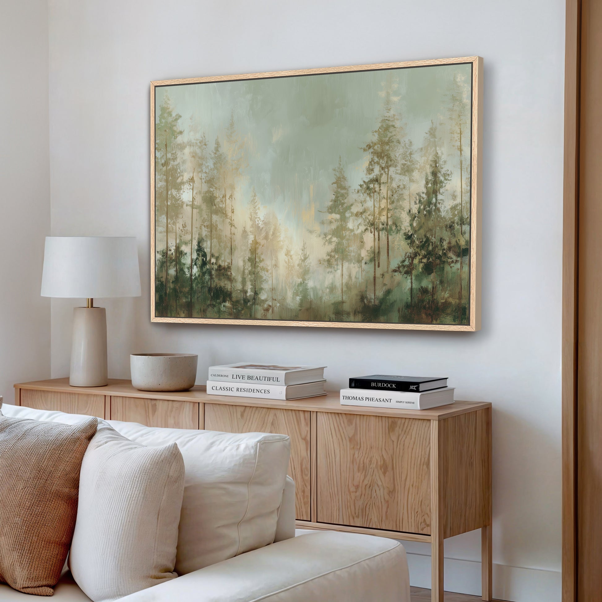a living room with a large painting of a forest scene hanging on the wall above a wooden cabinet. The cabinet has a bowl and a stack of books on top. There is a white couch in the room, and the overall color scheme is neutral, with the painting and the wooden cabinet providing a warm contrast.