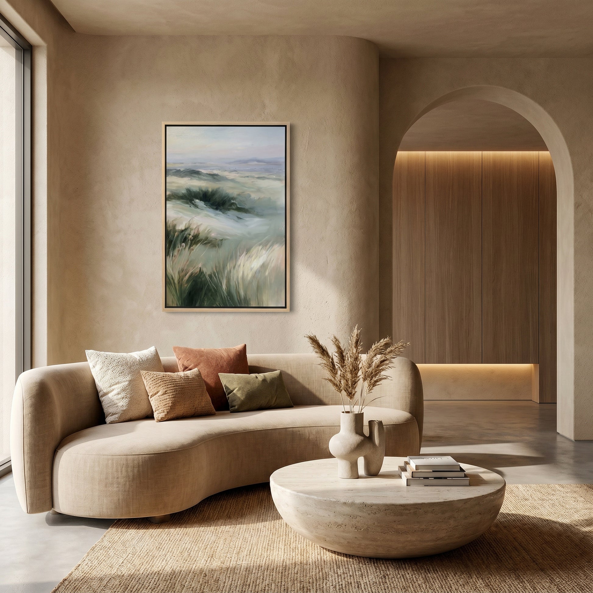 a modern living room with a large, curved sofa adorned with throw pillows and a vase of dried flowers. A framed landscape painting hangs on the wall above the sofa, and a round coffee table sits in front of it. The room features a wooden archway and a large window, creating a warm and inviting atmosphere.