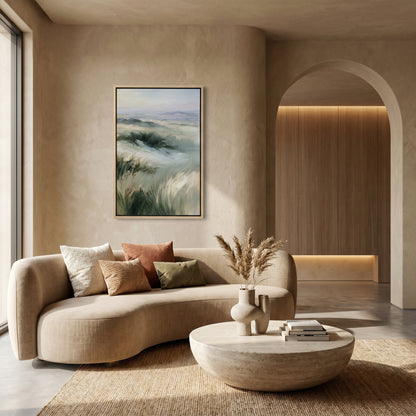 a modern living room with a large, curved sofa adorned with throw pillows and a vase of dried flowers. A framed landscape painting hangs on the wall above the sofa, and a round coffee table sits in front of it. The room features a wooden archway and a large window, creating a warm and inviting atmosphere.