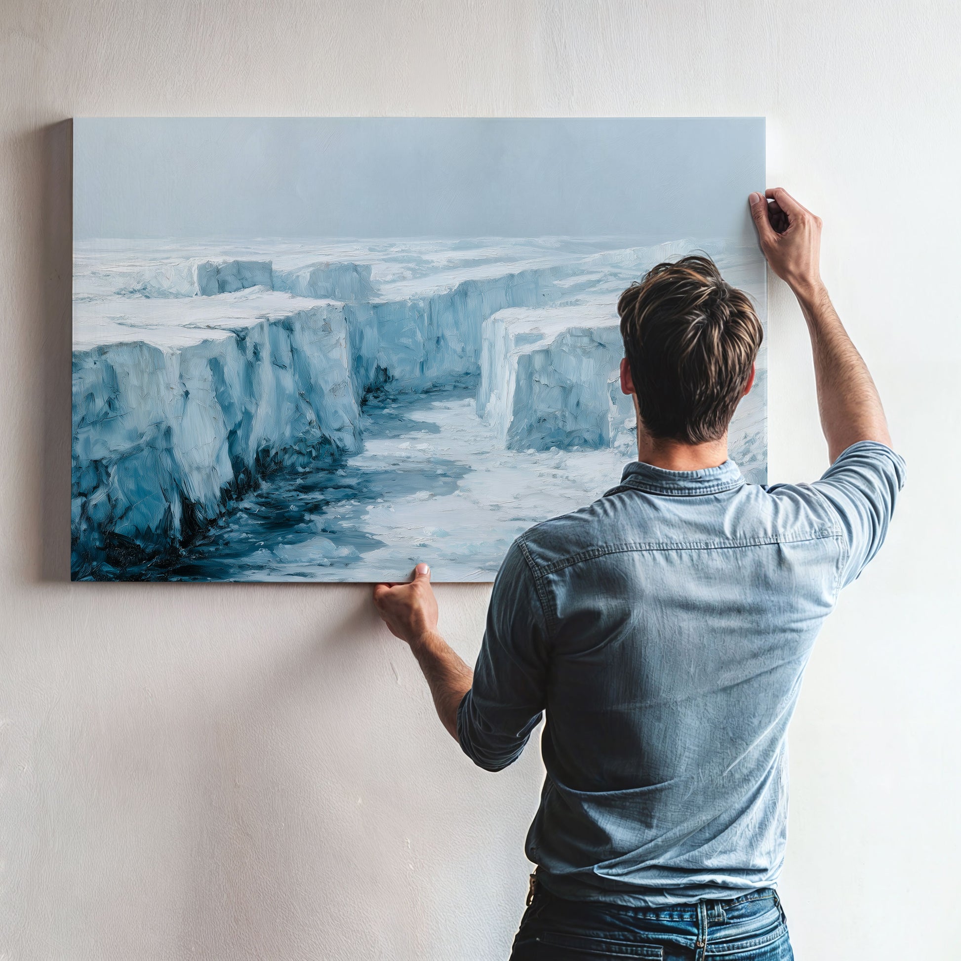 A man is hanging a large painting on a wall, which depicts a snowy, icy landscape.