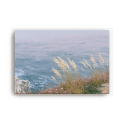 A serene landscape featuring a body of water with tall grasses swaying in the foreground, set against a hazy, blue sky.