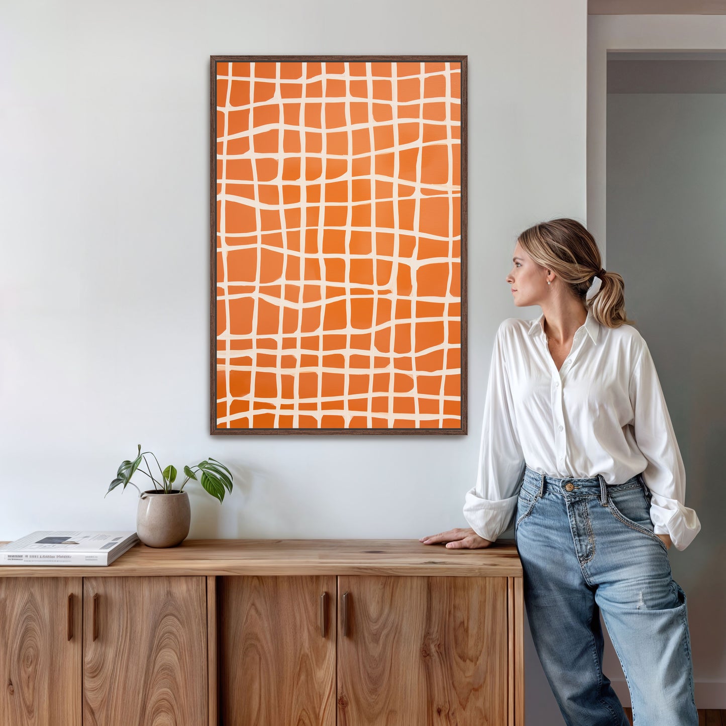 A woman in a white shirt and blue jeans stands next to a wooden cabinet, leaning against it with her hand on her hip. Above her, there is a framed orange and white abstract artwork on the wall.