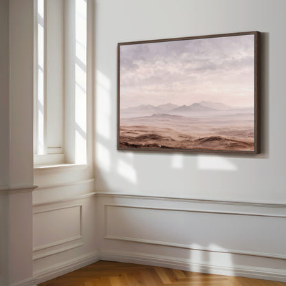 a large, framed painting of a desert landscape hanging on a white wall in a room with a window.