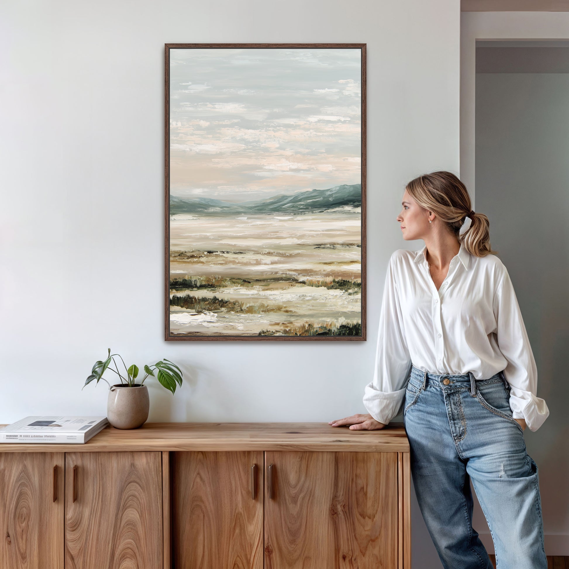 A woman in a white blouse and blue jeans stands next to a wooden cabinet, leaning against it and looking to the side. Above her, a framed landscape painting hangs on the wall.