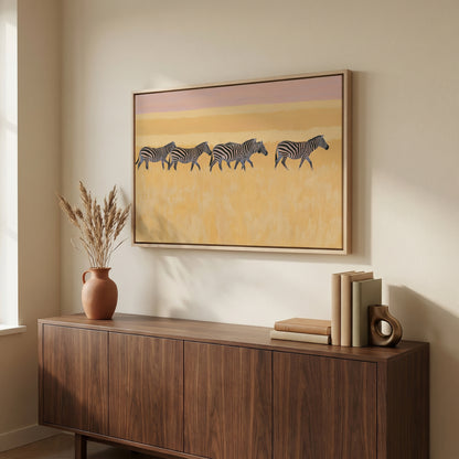 a wooden cabinet with a framed painting of zebras on the wall above it. The painting is a vibrant depiction of zebras in a field, with a warm yellow and orange background. The cabinet is situated next to a window, and there are some books and a vase on the cabinet.