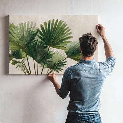 A man is hanging a large painting of green palm leaves on a wall.