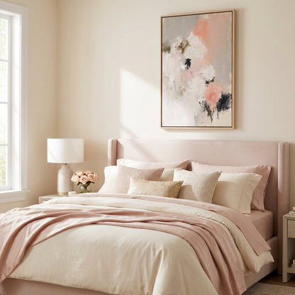 a serene and cozy bedroom with a large bed adorned with a pink and white bedspread and pillows. A framed abstract painting hangs above the bed, adding an artistic touch to the space. A lamp is placed on a side table next to the bed, providing additional lighting. The room has a window that allows natural light to enter, creating a warm and inviting atmosphere.
