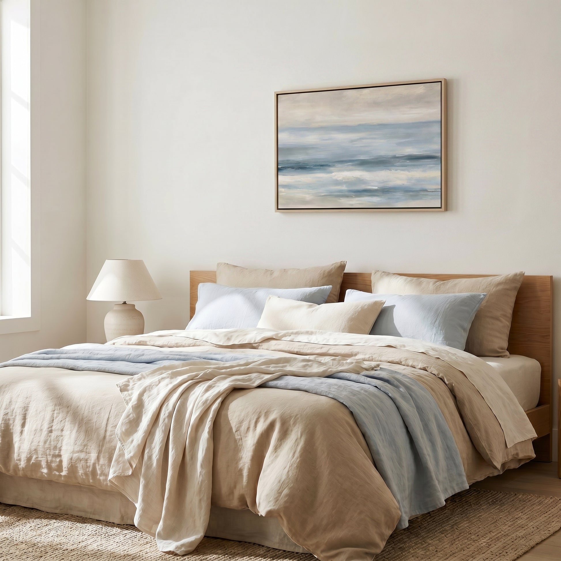 a serene and cozy bedroom with a large bed adorned with a beige comforter and blue throw blanket. Above the bed, a framed painting of a tranquil ocean scene hangs on the wall, adding a touch of nature to the room. The room is well-lit, with a lamp on a side table providing soft lighting, creating a warm and inviting atmosphere.