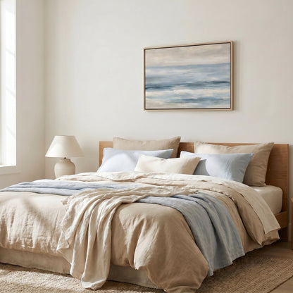 a serene and cozy bedroom with a large bed adorned with a beige comforter and blue throw blanket. Above the bed, a framed painting of a tranquil ocean scene hangs on the wall, adding a touch of nature to the room. The room is well-lit, with a lamp on a side table providing soft lighting, creating a warm and inviting atmosphere.