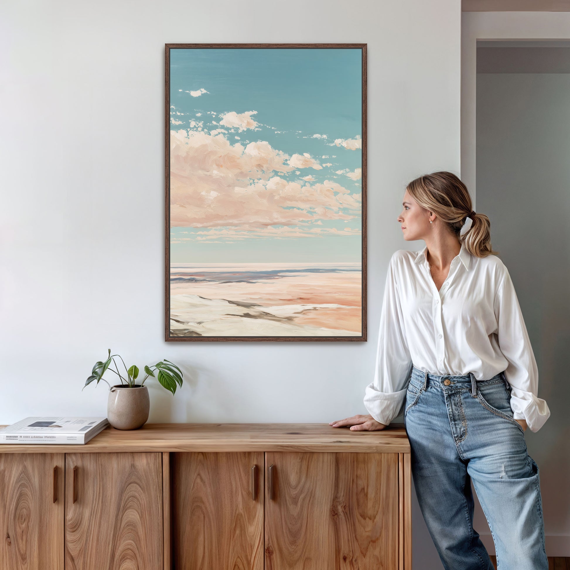 A woman in a white blouse and blue jeans stands in a room, leaning against a wooden cabinet, with a framed landscape painting hanging on the wall above her.