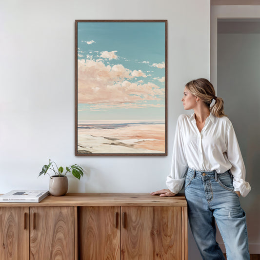 A woman in a white blouse and blue jeans stands in a room, leaning against a wooden cabinet, with a framed landscape painting hanging on the wall above her.