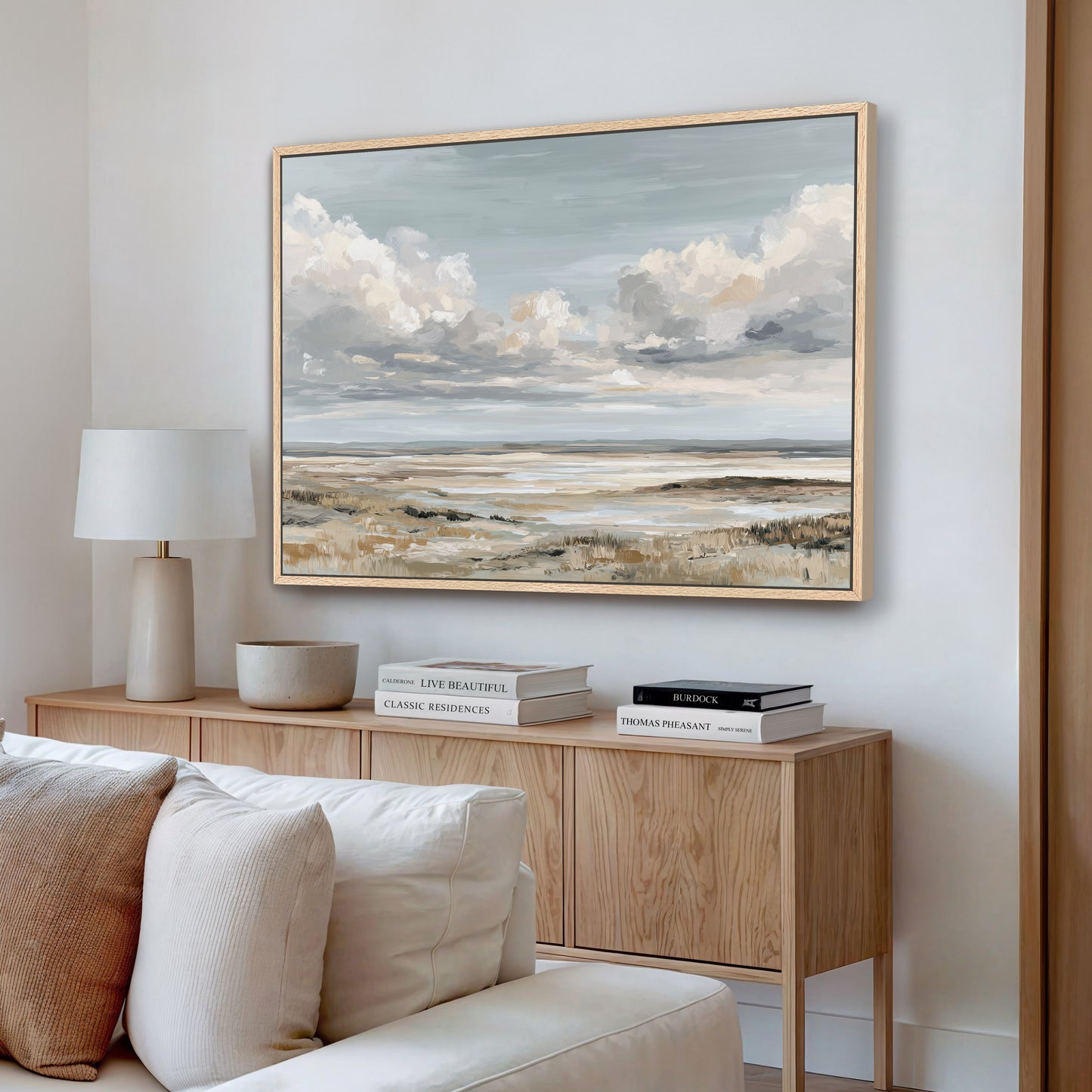a living room with a large painting of a landscape hanging on the wall above a wooden console table. The console table has a lamp, a bowl, and a stack of books on it. The couch is positioned in front of the console table, and the overall scene has a calm and serene atmosphere.