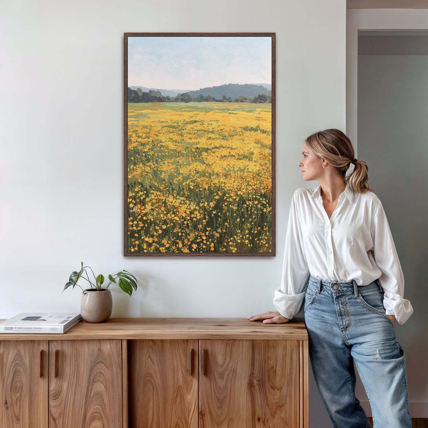 A woman stands in a room, leaning against a wooden cabinet, with a framed painting of a field of yellow flowers hanging on the wall above her.