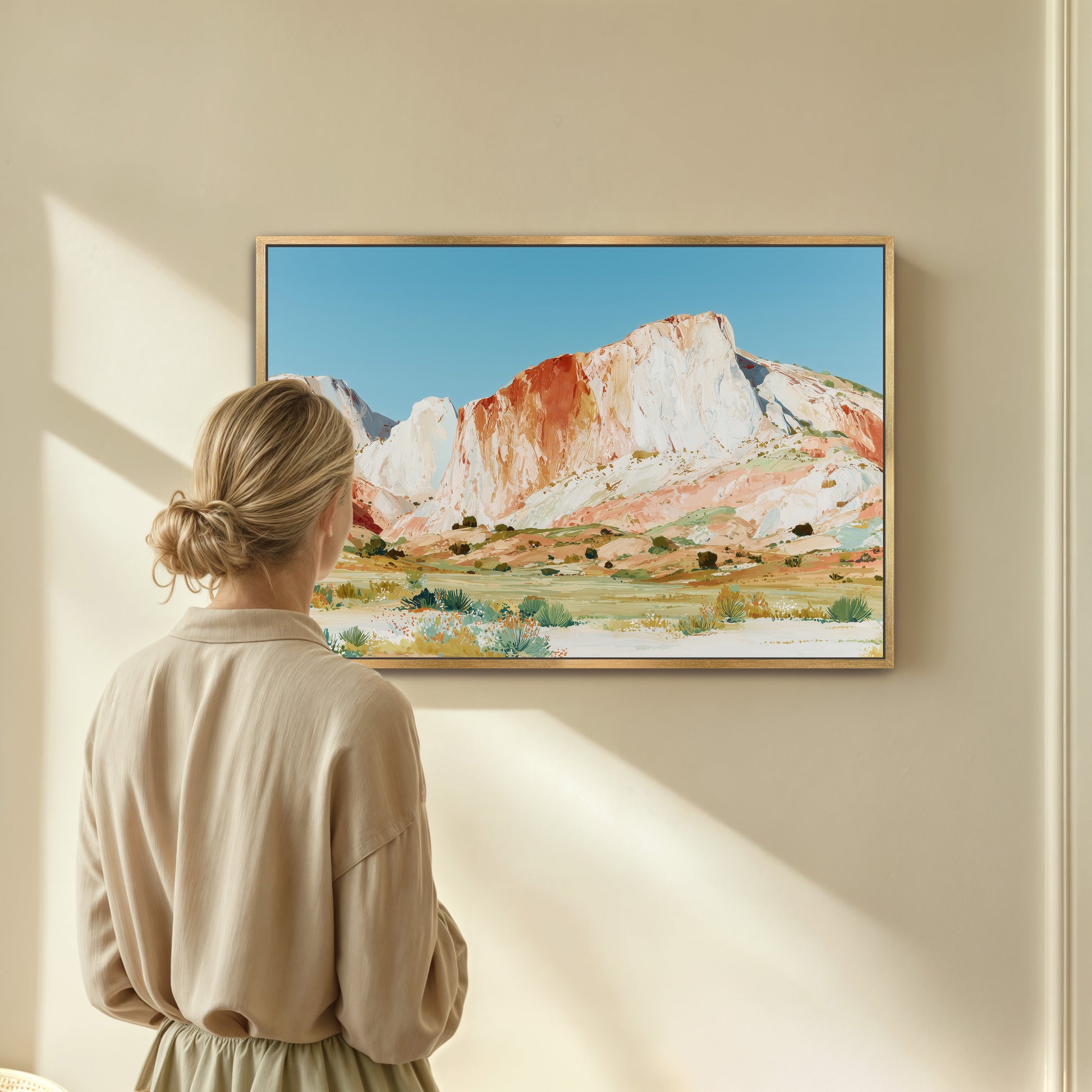 A woman is standing in front of a framed painting of a mountain landscape, looking at it with interest.