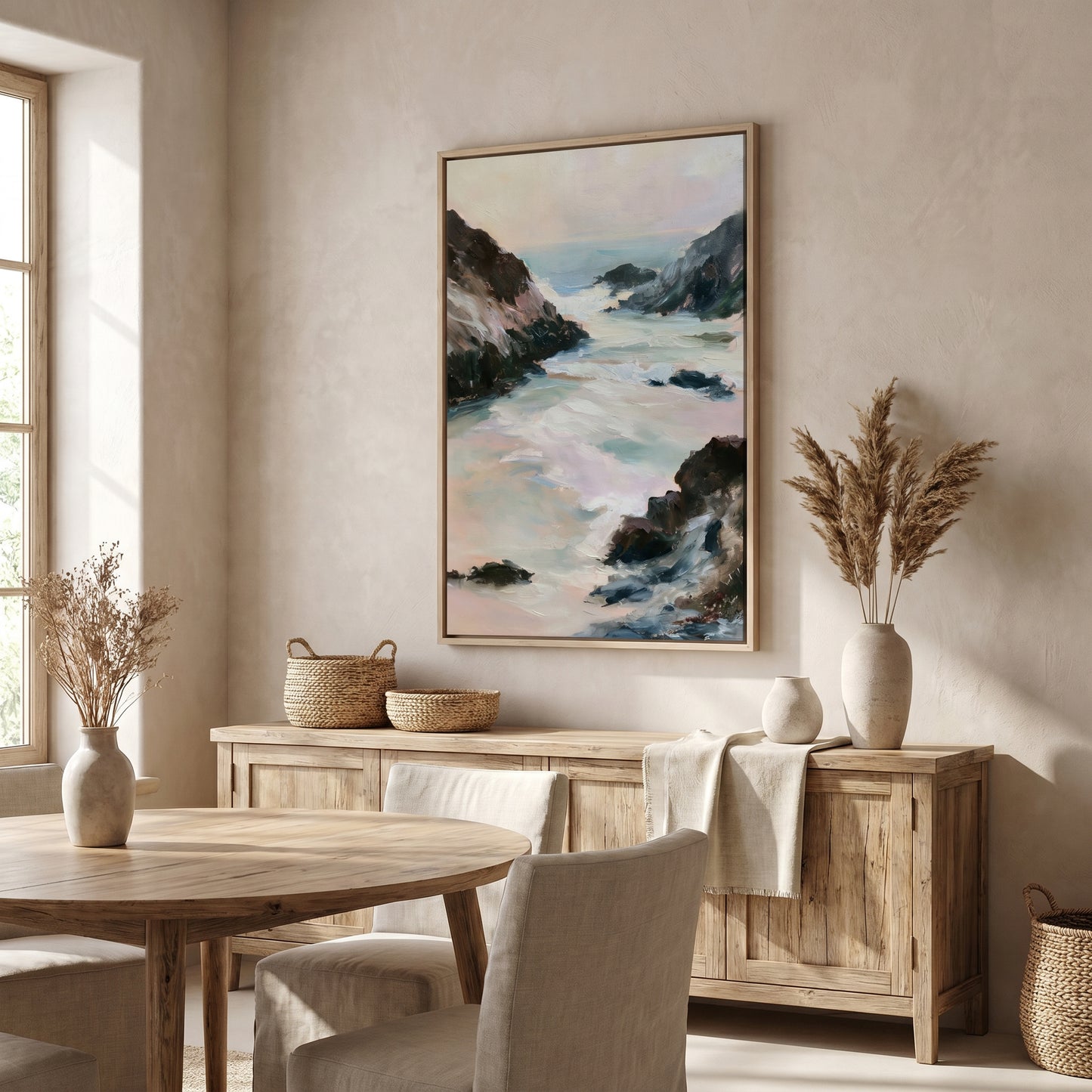 a serene dining room with a large painting of a rocky coastline hanging on the wall above a wooden table and chairs. The room has a neutral color palette, with a large window providing natural light and a view of the outdoors. The dining table is set with a vase of dried flowers and a woven basket, adding a touch of nature and rustic charm to the space.